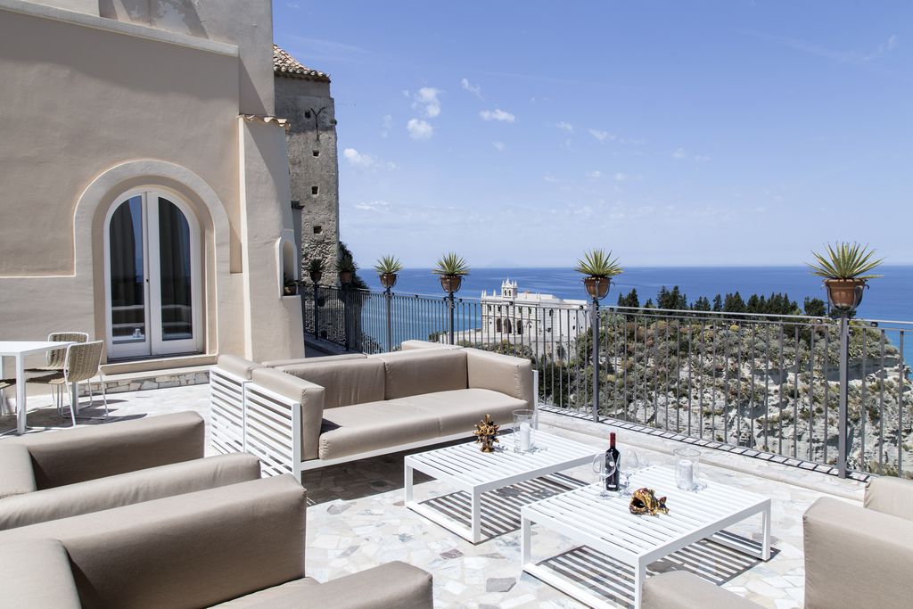 Exterior view of Dolce Vita a Tropea, Tropea, Vibo Valentia, Calabria, Italy. White building, terrace with beige and white sofas and tables, view overlooking the sea.